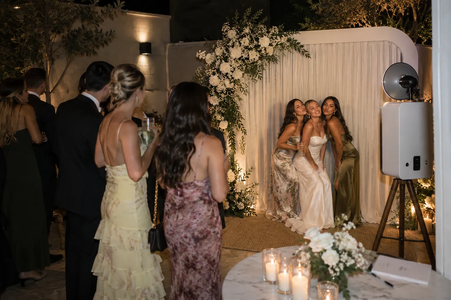 guests in evening wear posing at a glam photo booth with a draped white backdrop and roses